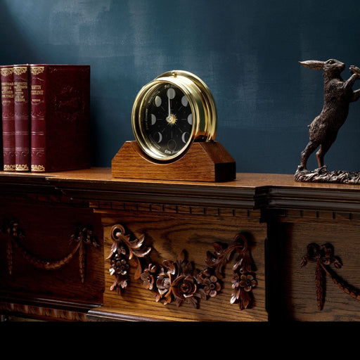 Weather Scientific Tabic Clocks Prestige Brass Moon Phase Clock With a Jet Black Dial Mounted on a Solid English Oak Mantel/Display Mount displayed on table